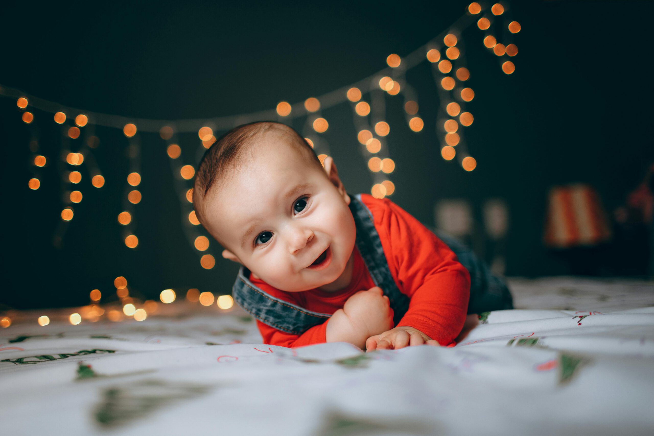 Cute baby boy lying on bed with festive string lights creating a warm, cozy atmosphere.